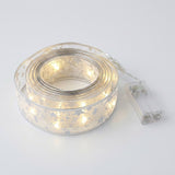 Coiled silver patterned ribbon light string glows warm white beside a connected clear battery box on a pale gray background.
