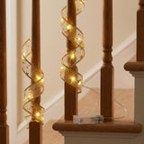 Gold patterned ribbon light string spirals around wooden staircase spindles, glowing warm white beside a clear battery box on the ledge.