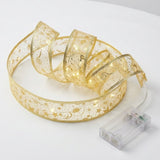 Gold patterned ribbon light string forms loose glowing loops beside an empty clear battery box on a white surface.