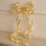 Sheer gold ribbon light string forms a glowing bow against a cream wall, with patterned tails curling onto a wooden surface.