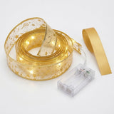 Coiled gold patterned ribbon light string glows warm white beside a clear battery box and plain gold ribbon strip on white.