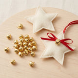 Gold-color jingle bells lie beside two cream felt stars on pale wood, with one bell attached to a red ribbon bow.