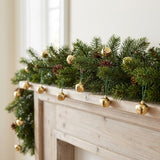 Gold-color jingle bells hang from green cords on an evergreen garland with pinecones across a white wooden mantel beside a window.