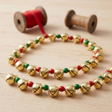 Gold jingle bells strung with red and green beads rest on a pale wooden tabletop beside ribbon and thread spools.