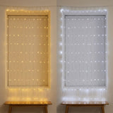 Side-by-side windows with closed blinds display warm white curtain lights on the left and cool white lights on the right.