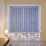 Cool white LED curtain string lights draped over a window with sheer curtains in a bright room, a remote resting on a wooden sideboard.