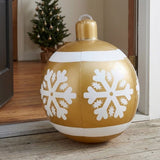 A gold inflatable Christmas ornament with white snowflakes and bands sits outside an open doorway with a decorated tree inside.