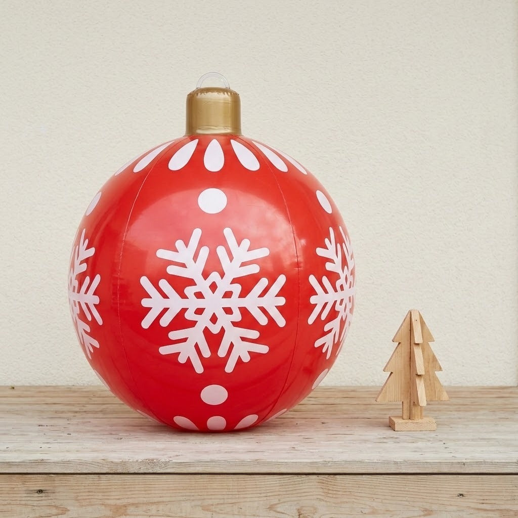 A red inflatable Christmas ornament with white snowflakes and a gold cap stands beside a wooden tree against a cream wall.