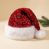A festive Santa hat with a red sequined crown and a fluffy white faux-fur brim and pom-pom, photographed on a warm beige background with a small green pine branch prop at the side.