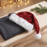 A festive Christmas Santa hat with red sequins and white faux fur trim, draped over a folded dark gray sweater on a light wooden table with warm string lights in the background.