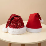 Two Santa hats displayed on a round cream table against a beige wall. One hat is made of red sequins with a white faux-fur trim and pom-pom, and the other is a solid red felt Santa hat with white trim and pom-pom.