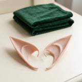 A pair of peach latex elf ears rests on a cream surface beside folded dark green fabric.
