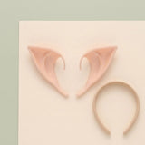 A pair of peach latex elf ears and a thin gold-colored headband lie on a cream board against pale green.
