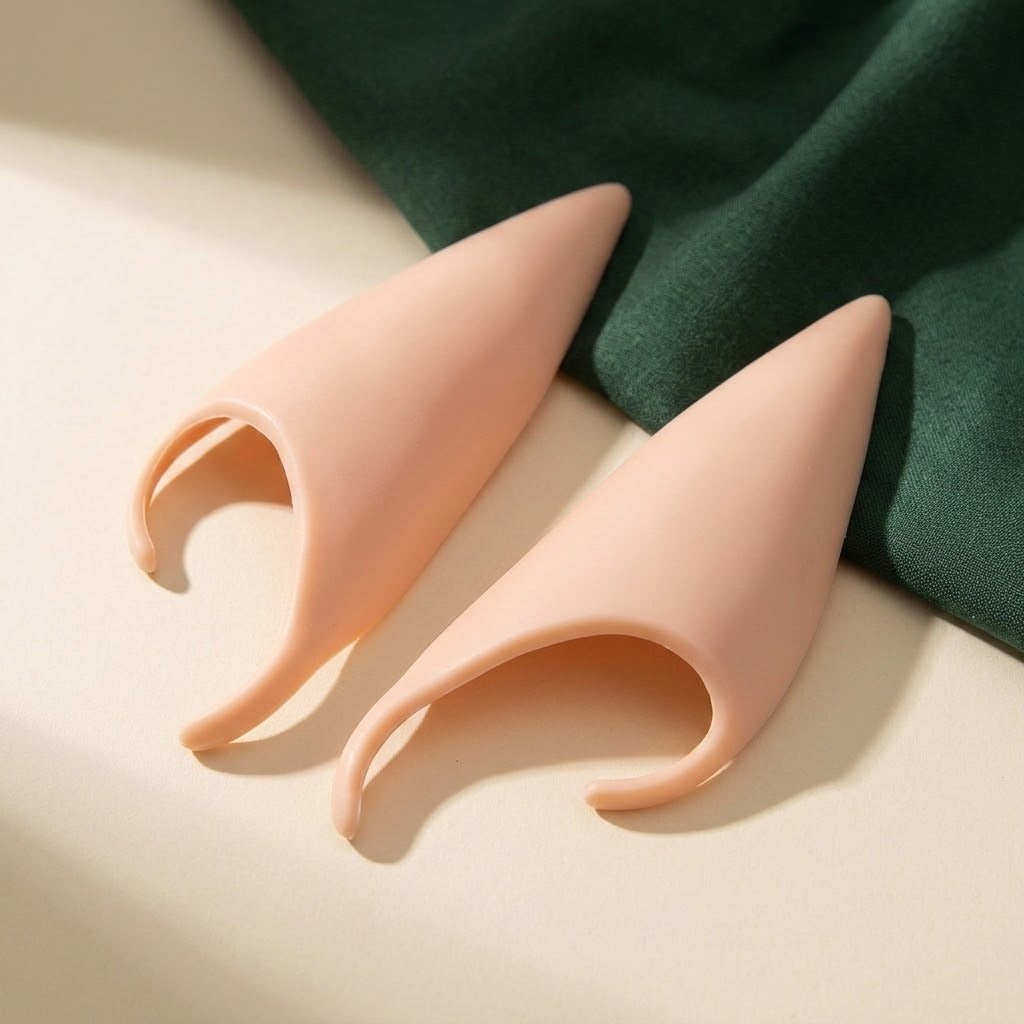 Two peach latex elf ears display their smooth outer surfaces and curved openings beside dark green fabric.