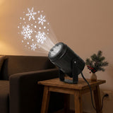 A black snowflake projector sits on a wooden side table beside a sofa, casting white snowflakes onto a beige wall.