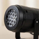 The black snowflake projector has a textured front rim, clear circular lenses, and a side screw against a blurred beige background.
