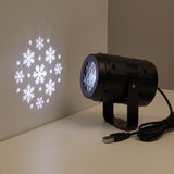 A black projector on a white tabletop casts white snowflakes onto a beige wall, with its USB plug and coiled cable visible.