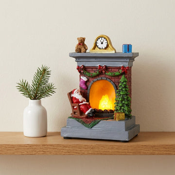 A Santa fireplace Christmas decorative light with a gray base glows on a wooden shelf beside a white vase of evergreen sprigs.