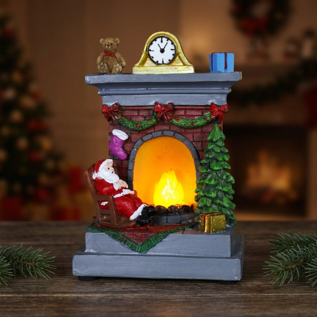 A Santa fireplace Christmas decorative light glows orange on dark wood between evergreen sprigs, with a blurred tree and fireplace behind.