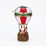 A red and white hot air balloon Christmas decorative light with gold trim carries Santa in a brown basket against white.