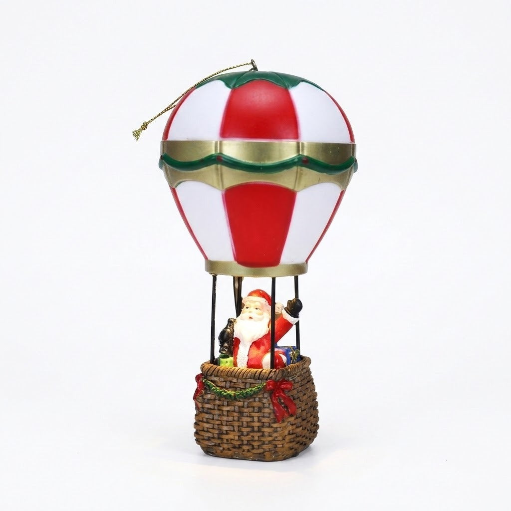 A red and white hot air balloon Christmas decorative light with gold trim carries Santa in a brown basket against white.