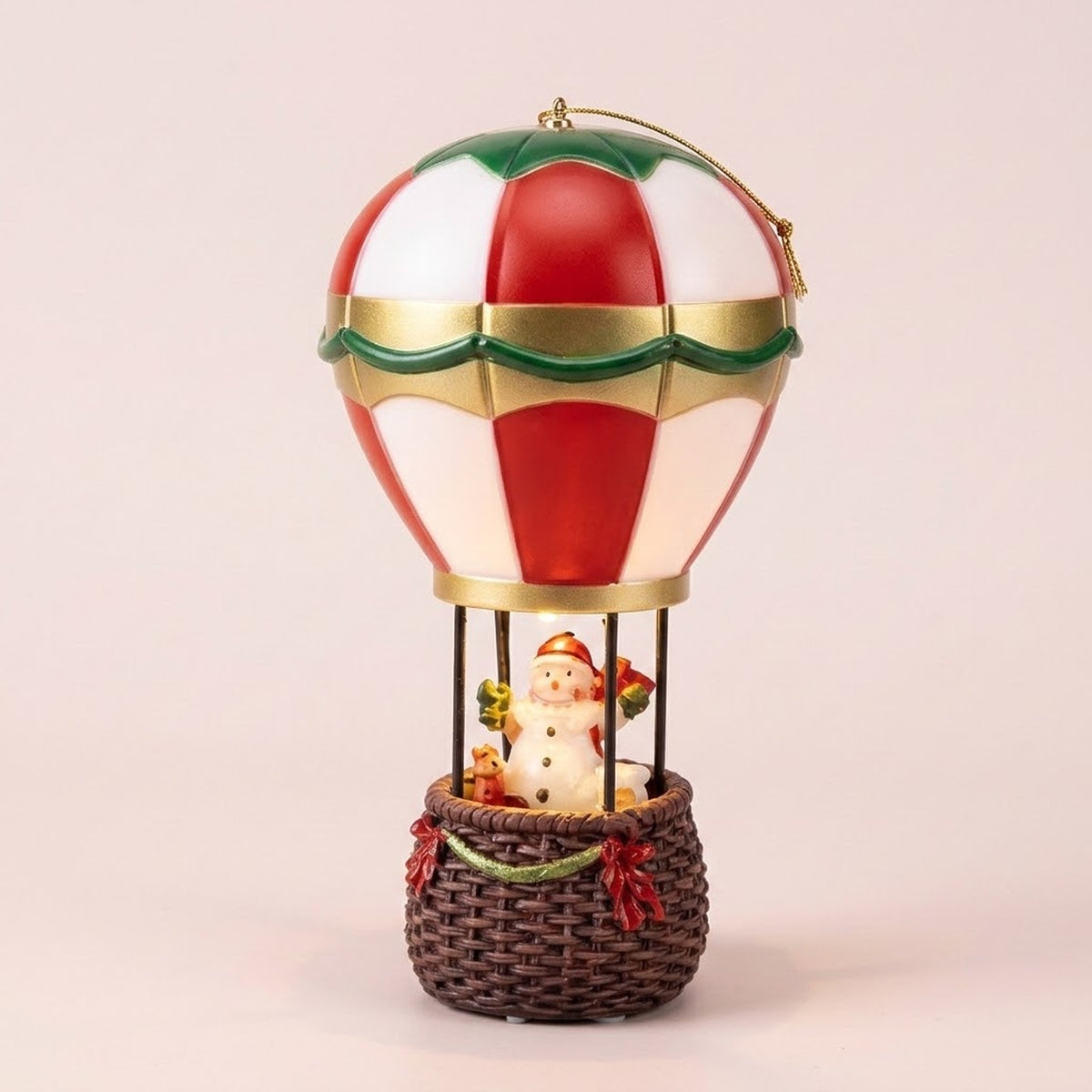 Snowman hot air balloon Christmas decorative light has red and white panels, gold and green trim, and a brown basket against pale pink.