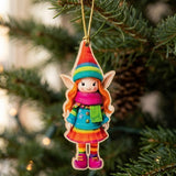 A flat wooden Christmas ornament shaped like an orange-haired elf in rainbow clothing hangs by a gold string from an evergreen branch.