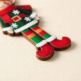 A flat wooden elf Christmas ornament rests on a cream surface, showing brown edges, striped legs and red boots in close-up.