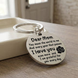 Silver keychain with a circular Dear Mom message pendant and rose motif leans against a cream tray on a wooden surface.