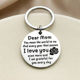 Silver circular Dear Mom keychain with a black message and rose motif rests on a rounded cream surface.