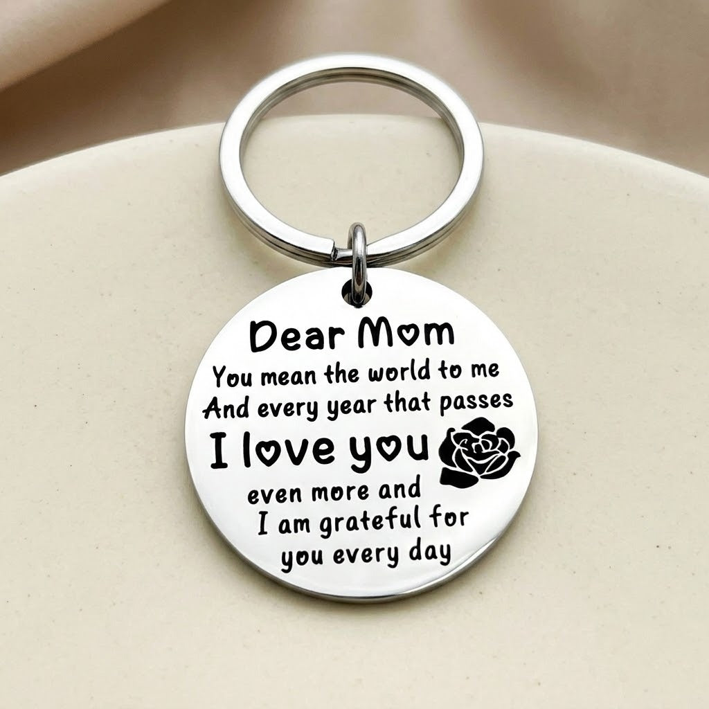 Silver circular Dear Mom keychain with a black message and rose motif rests on a rounded cream surface.