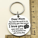 Silver Dear Mom keychain lies beside perpendicular wooden rulers with centimeter markings on a cream background.