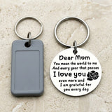 Round silver keychain with a Dear Mom message and black rose beside a gray rectangular key tag on a beige stone surface.