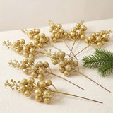 Gold glitter berry picks lie across a cream textured surface beside a green evergreen sprig.