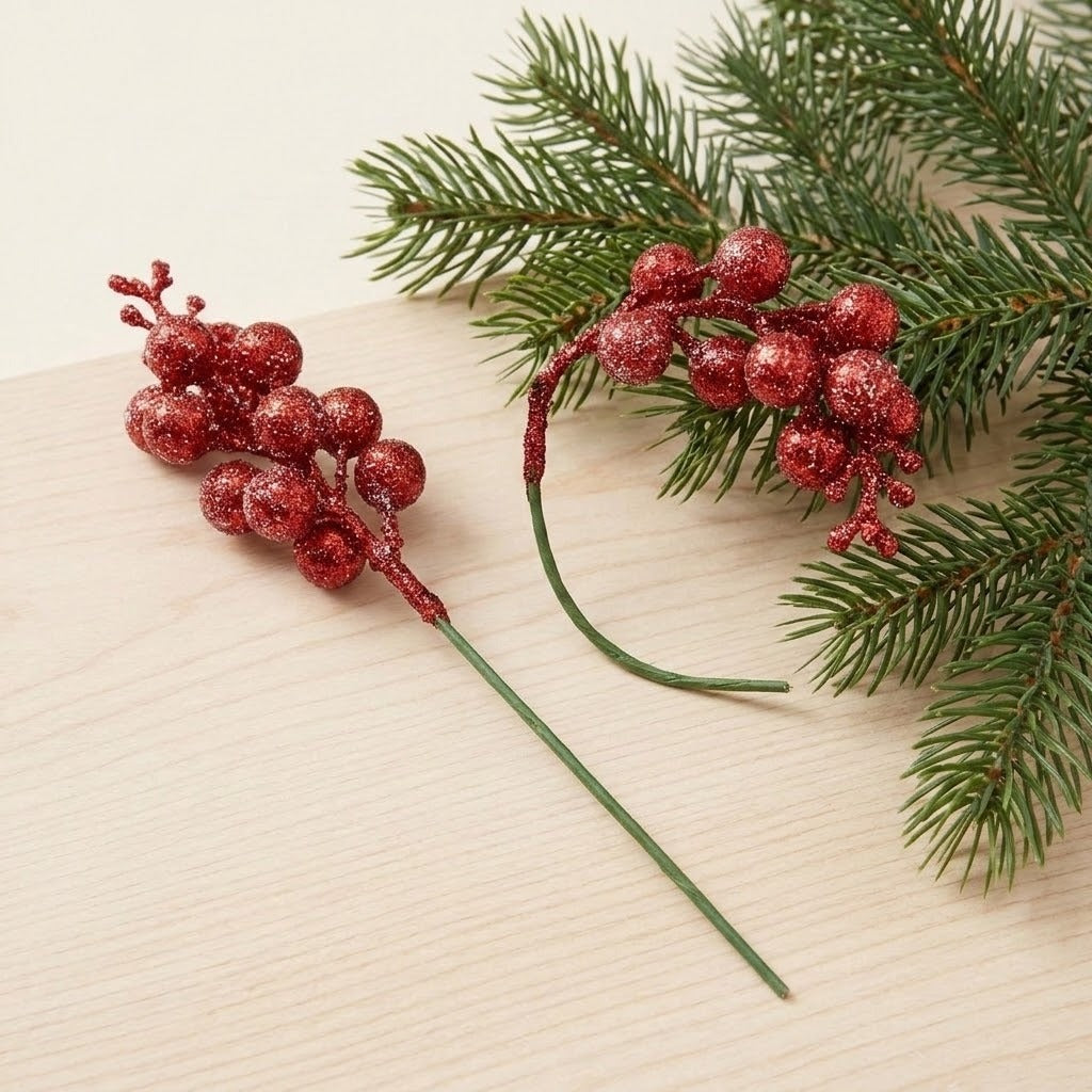 Two red glitter berry picks, one straight and one curved, rest on pale wood beside green evergreen branches.
