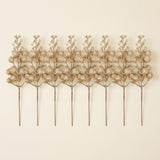 Eight champagne gold glitter berry picks with long slender stems lie side by side on a cream background.