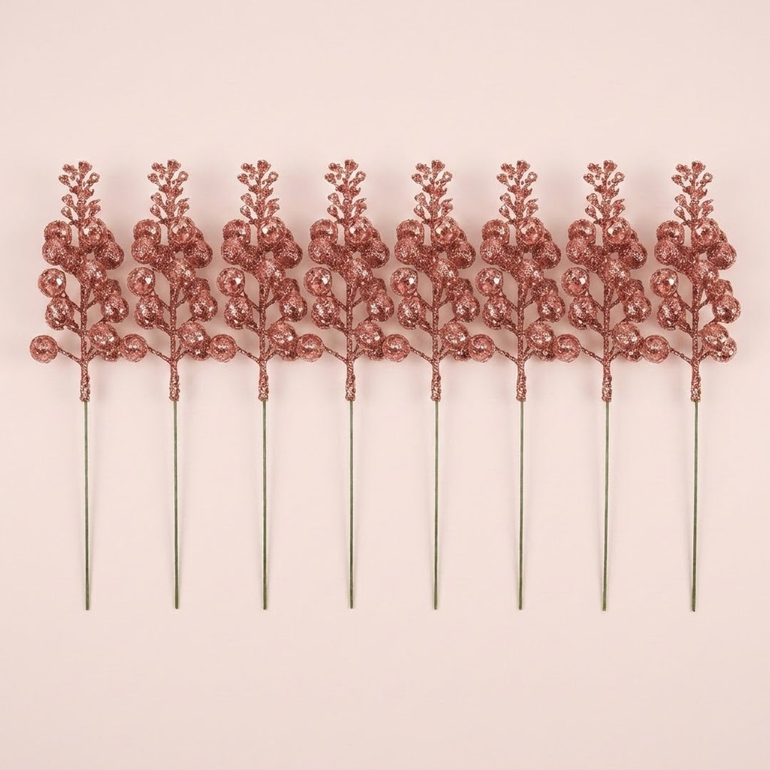 Eight rose gold glitter berry picks with long slender stems lie in a row on a pale pink background.