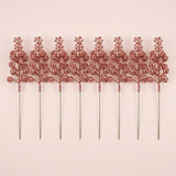 Eight rose gold glitter berry picks with long slender stems lie in a row on a pale pink background.