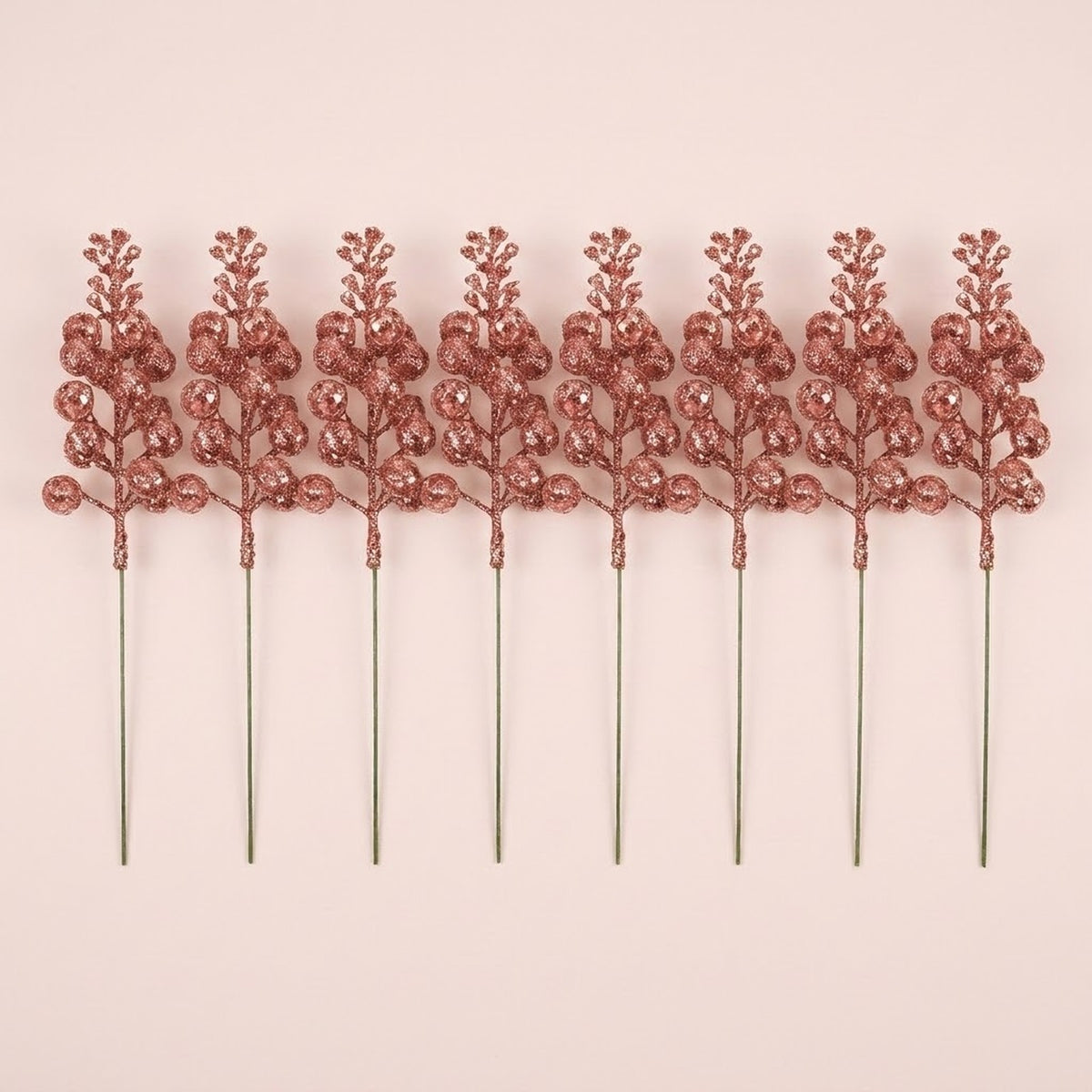 Eight rose gold glitter berry picks with long slender stems lie in a row on a pale pink background.