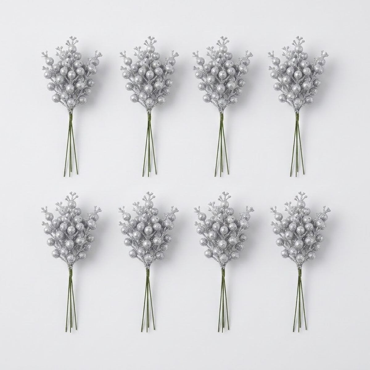 Eight bunches of silver glitter berry picks with green stems form two rows of four on a pale gray background.