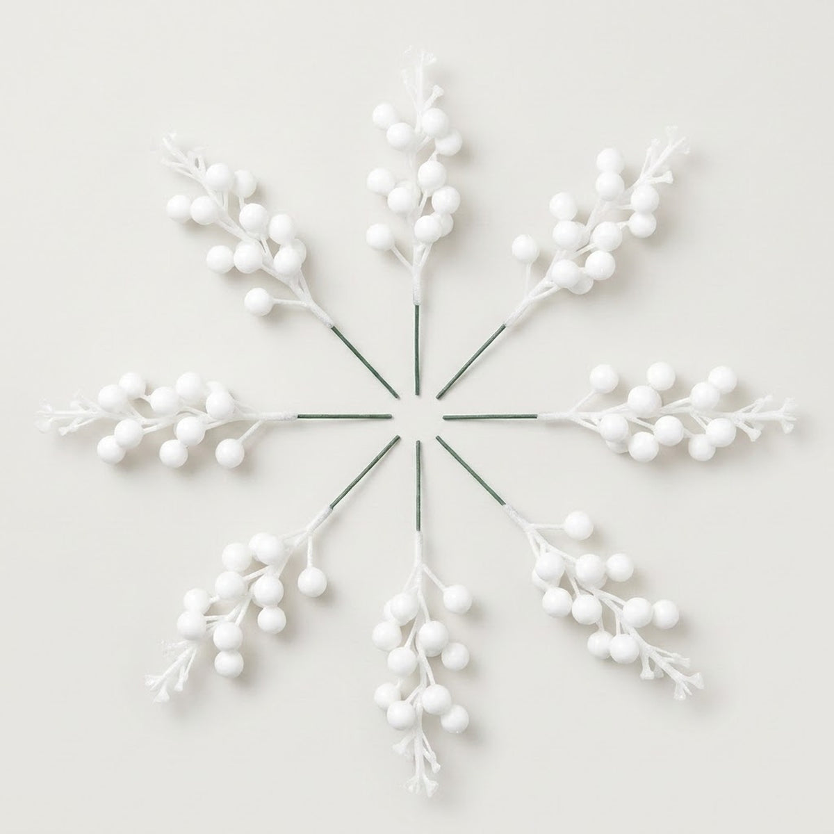Eight matte white berry picks with green stems form a radial arrangement on an off-white background.