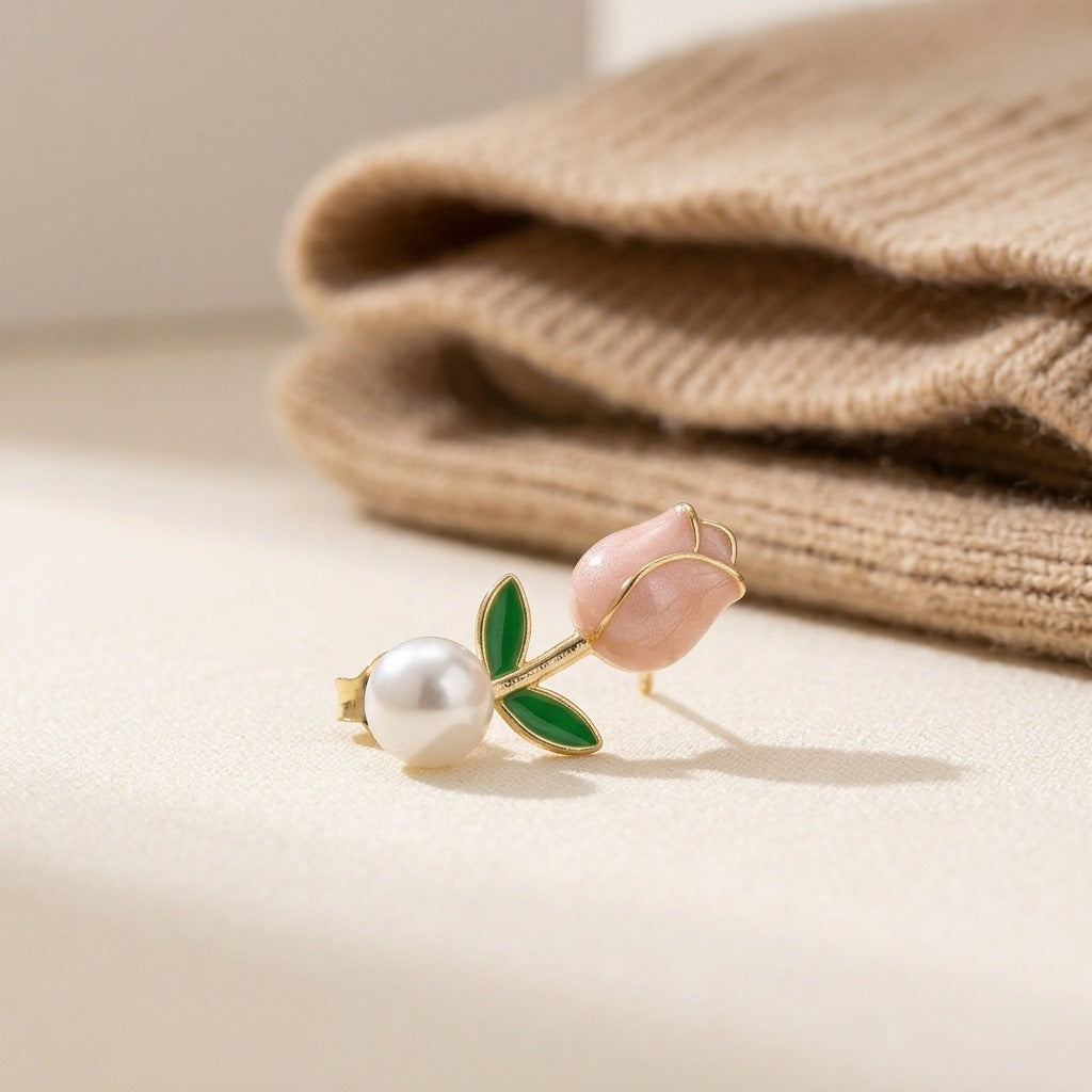 A gold-tone tulip stud earring with a pink bud, green leaves, and white pearl-like accent rests beside folded beige knit fabric.