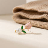 A gold-tone tulip stud earring with a pink bud, green leaves, and white pearl-like accent rests beside folded beige knit fabric.