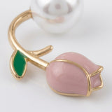 A close-up of a tulip stud earring shows gold-tone edging, a pink bud, green leaf, and cropped pearl-like bead against white.