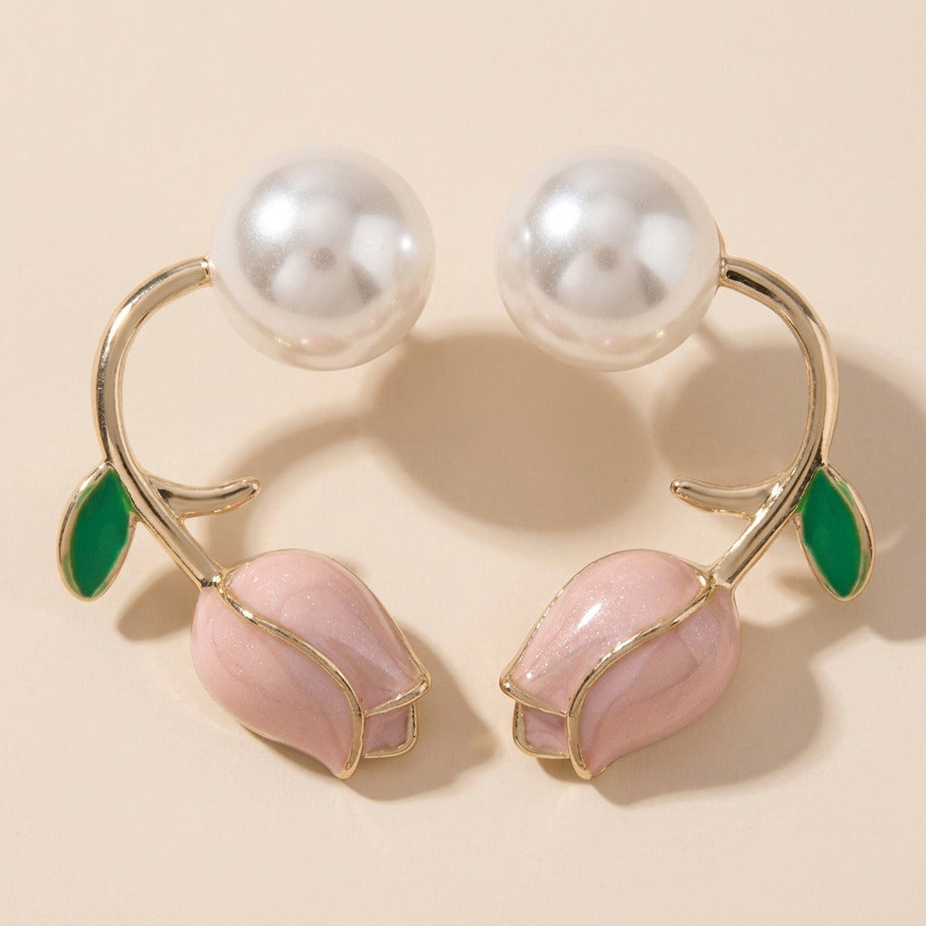 Two mirrored gold-tone tulip stud earrings rest on beige, featuring pink buds, green leaves, and white pearl-like beads with pronounced shadows.