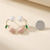 Pink tulip stud earrings with gold-tone stems, green leaves, and white pearl-like beads sit beside a silver-colored disc on beige.