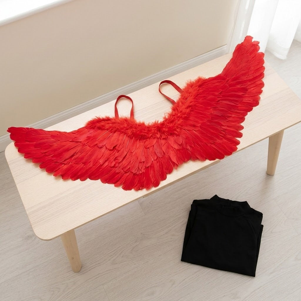 Red feather costume wings with matching straps rest on a pale wooden bench beside a folded black garment on the floor.
