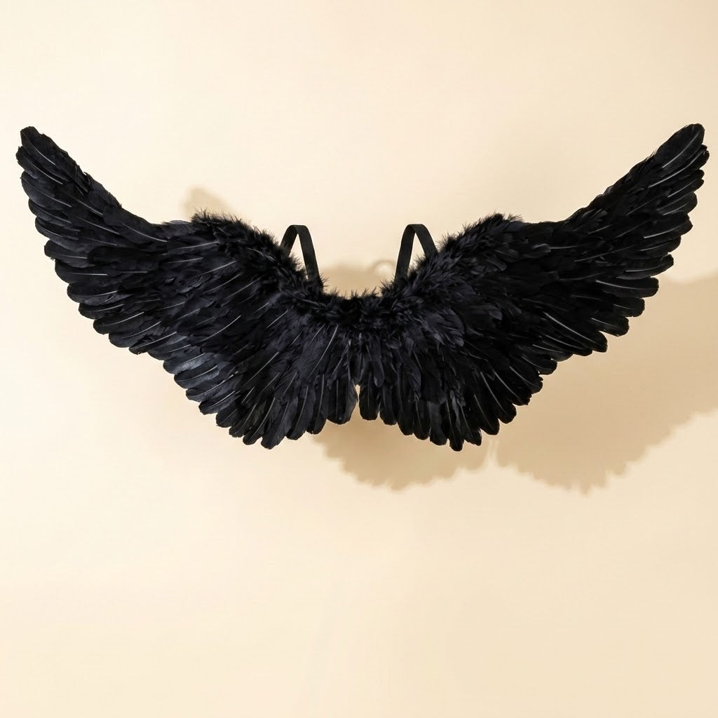 Black feather costume wings with visible shoulder straps are displayed against a cream background, casting a soft shadow.