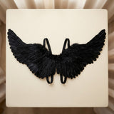 Black feather costume wings with looped elastic straps lie flat on a cream tabletop beside beige curtains.