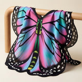 Pink and turquoise chiffon butterfly wing cape draped over a light wooden rail against a warm beige background.